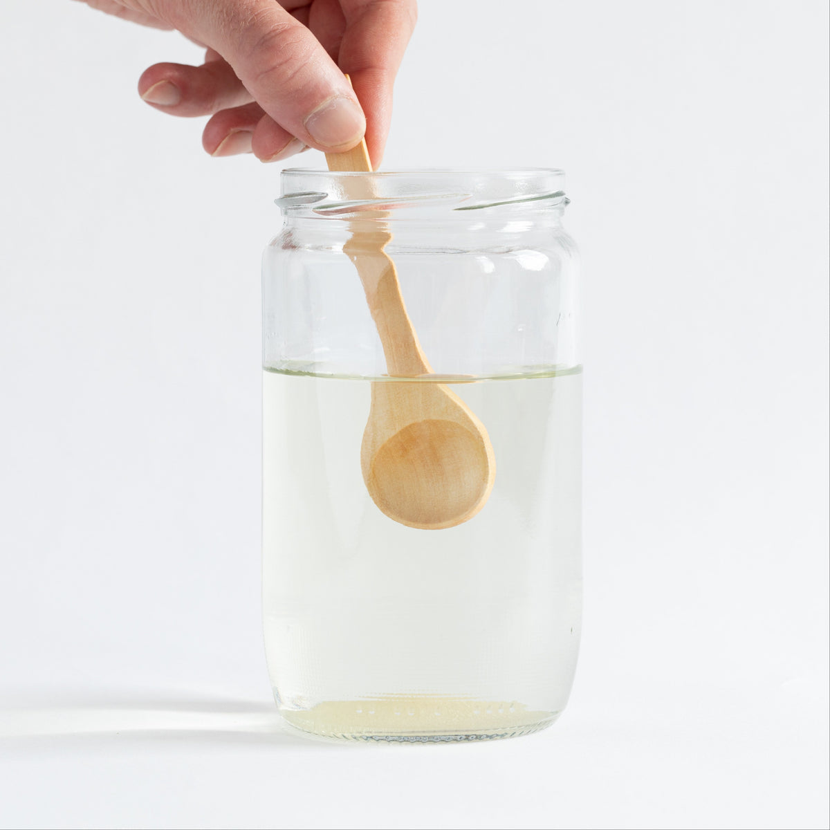 A hand holding a small light wood spoon above a clear glass jar filled with clear liquid, photographed on a clean white background.